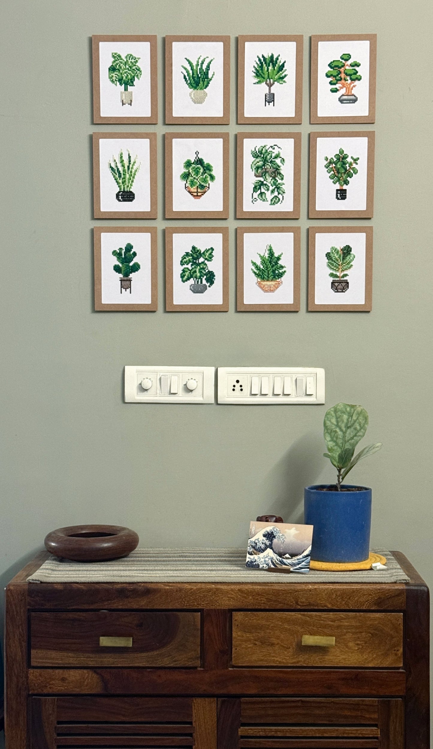Tweleve framed dimaond painting pictures on a wall above a wooden cabinet with decorative items.