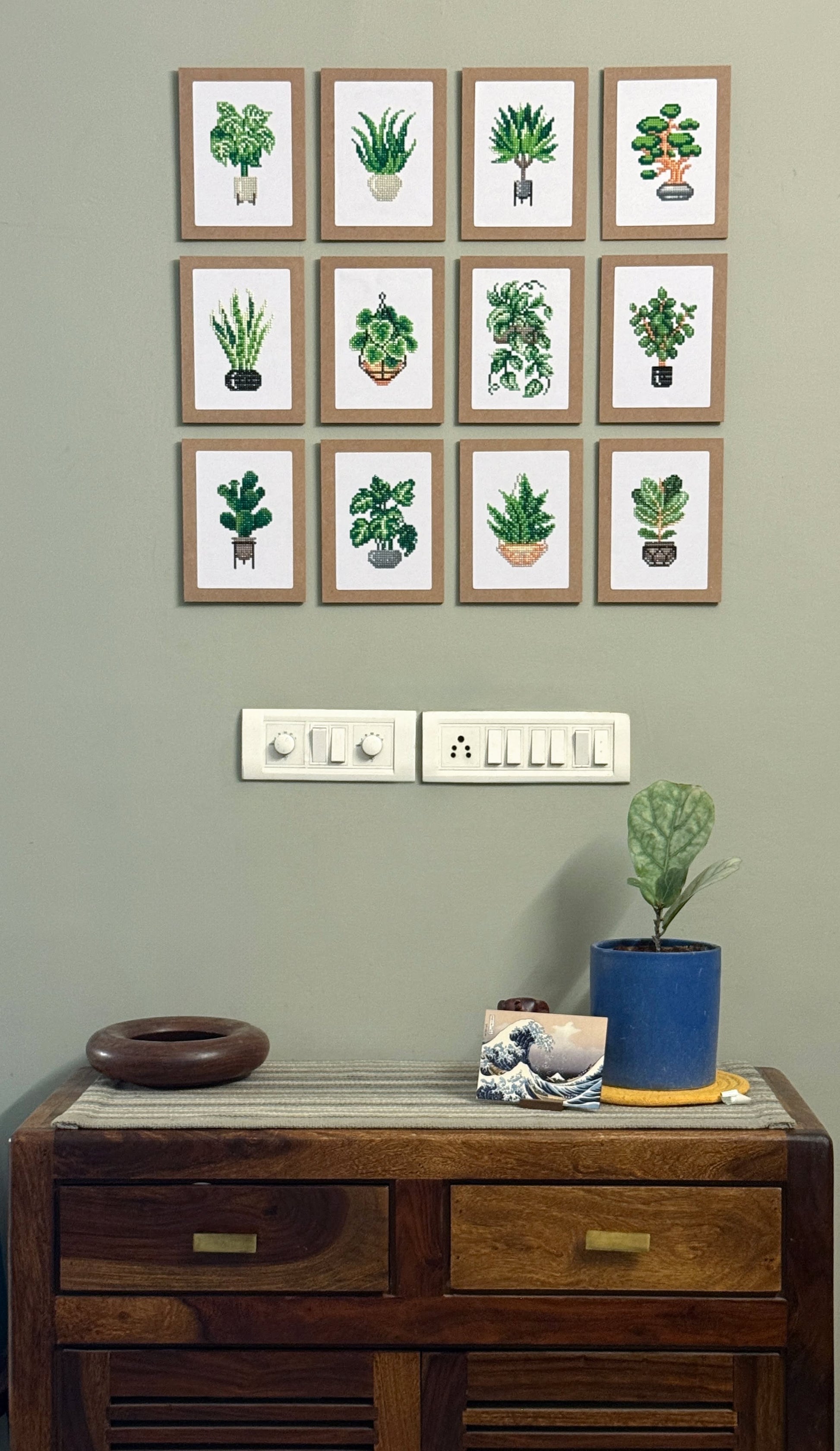 Tweleve framed dimaond painting pictures on a wall above a wooden cabinet with decorative items.